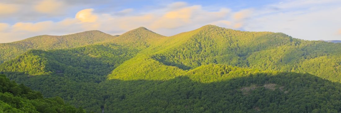 Warm evening light sweeping across overlapping green Blue Ridge summits below pastel clouds — Blue Ridge Parkway