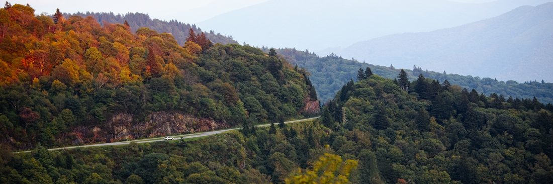 Lone car on a winding Blue Ridge Parkway road beside autumn ridges fading into hazy blue distance — Waterrock Knob, Blue Ridge Parkway